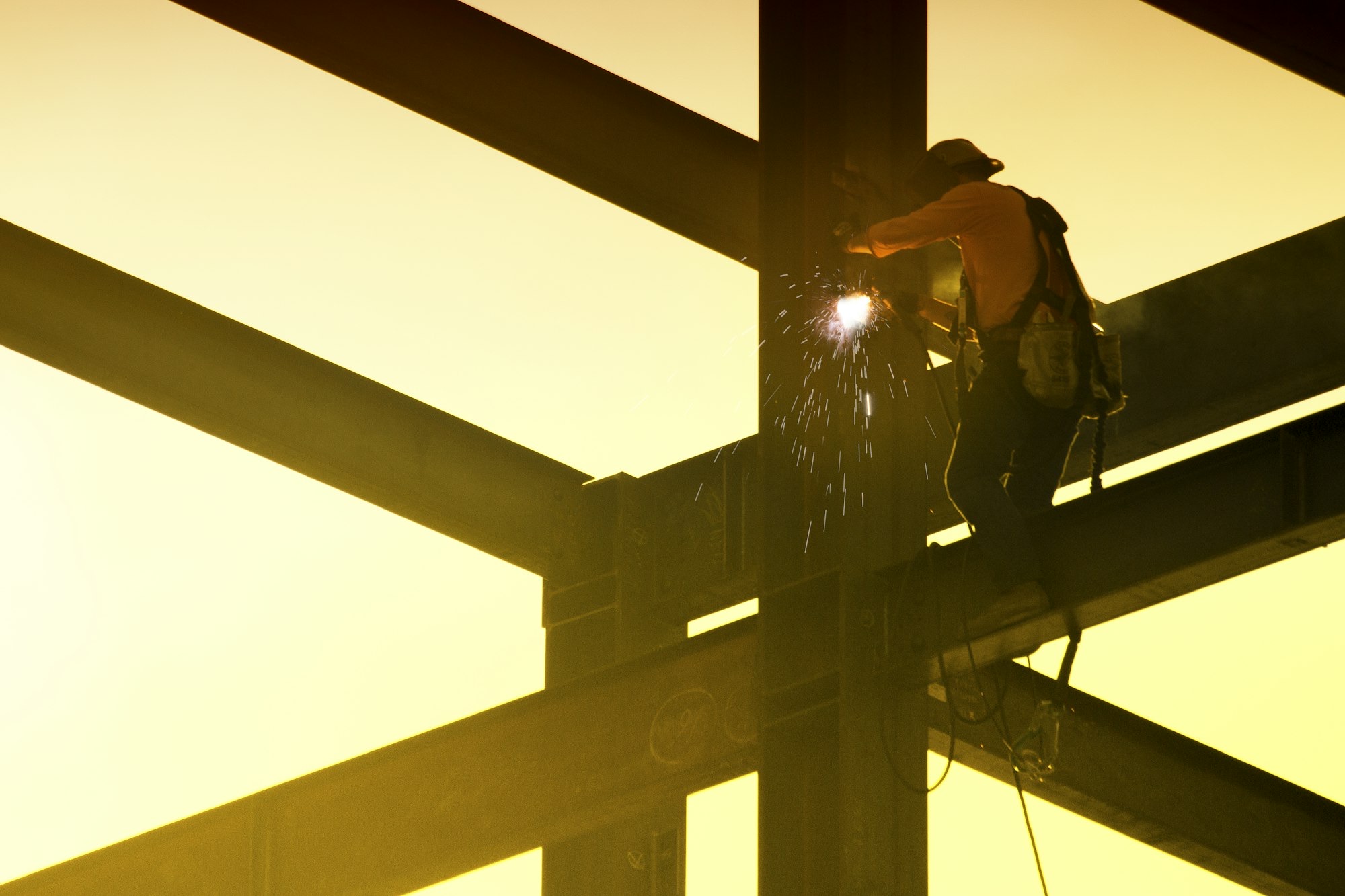 A steelworker welding high up on the beams against a golden sky.