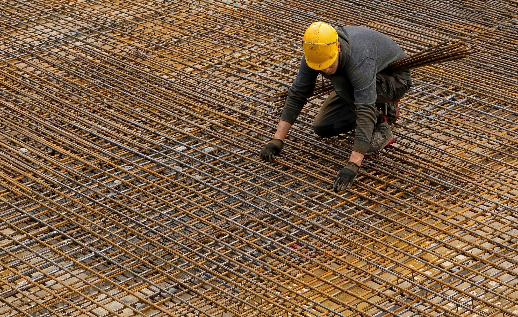 A worker in a hard hat and work jacket tying a rebar mat on site.
